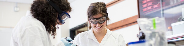 Two female student in scrubs and protective eyewear in a chemistry lab.